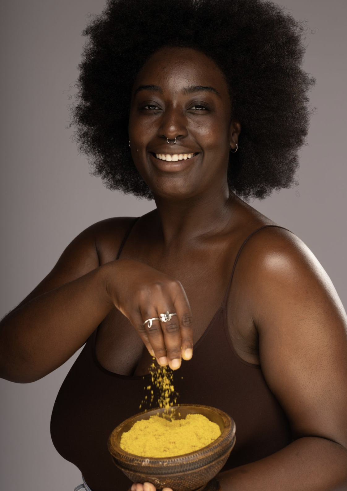 A person with a cheerful expression is pouring a yellowish organic scrub from a wooden bowl, representing the product 'Anyanwu' Hyperpigmentation Scrub, which is designed to exfoliate and brighten the skin.