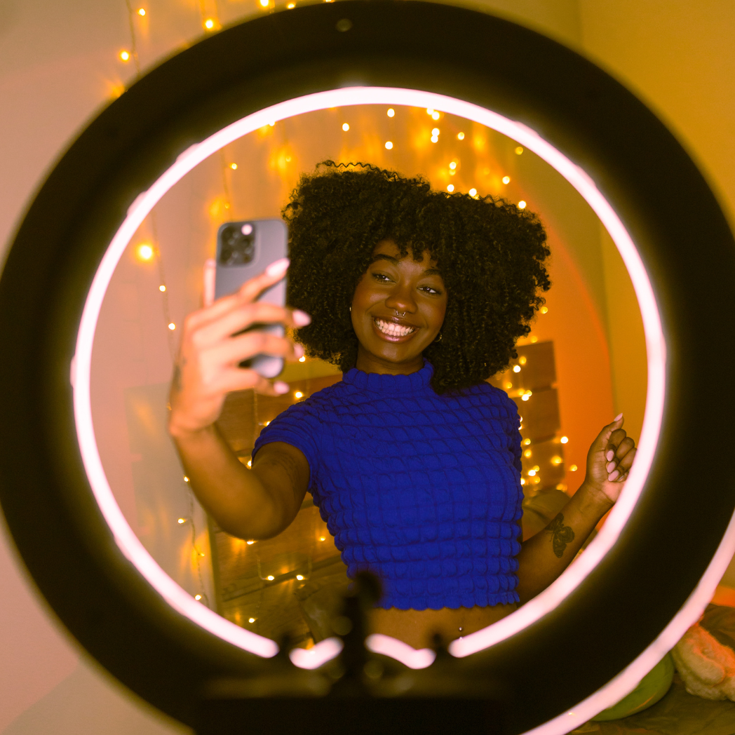 Person taking a mirror selfie with a ring light in a room decorated with string lights.