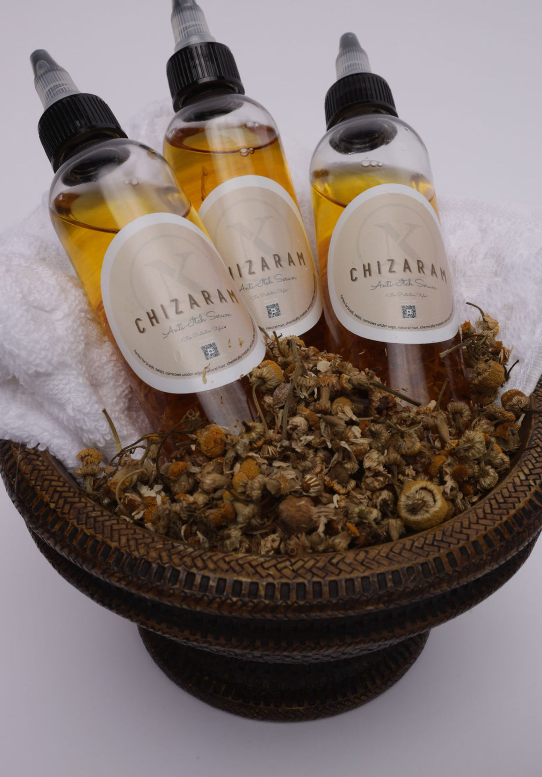 Three bottles of 'Chizaram' Anti-Itch Serum placed in a wooden basket with a towel and dry herbs surrounding them, indicating natural ingredients.
