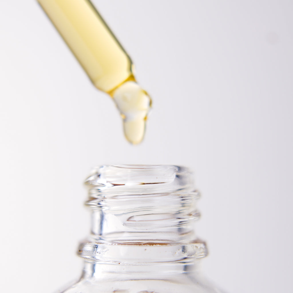 Close-up of a dropper with yellow liquid dripping into a clear bottle of "Ugomma" Facial Oil on a white background