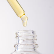 Close-up of a dropper with yellow liquid dripping into a clear bottle of "Ugomma" Facial Oil on a white background