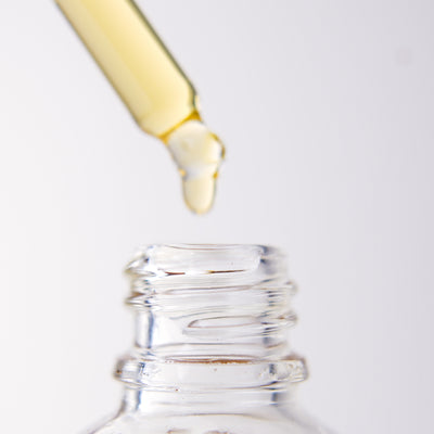 Close-up of a dropper with yellow liquid dripping into a clear bottle of "Ugomma" Facial Oil on a white background