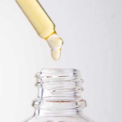 Close-up of a dropper with yellow liquid dripping into a clear bottle of "Ugomma" Facial Oil on a white background