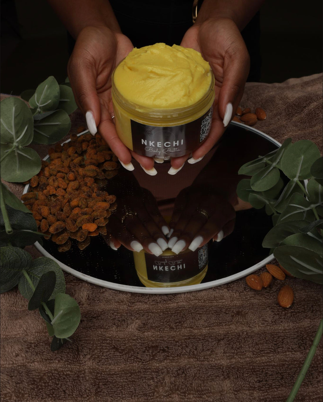 A jar of 'Nkechi' Body Butter held in a person's hands, with the product's name visible on the label. The background features a wooden surface with nuts and greenery.