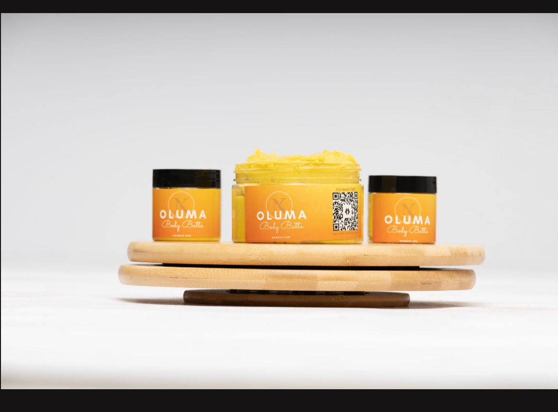 A jar of 'Oluma' Body Butter placed on a wooden board with two smaller jars to the sides. All have orange labels with the brand name 'Oluma' written in white.