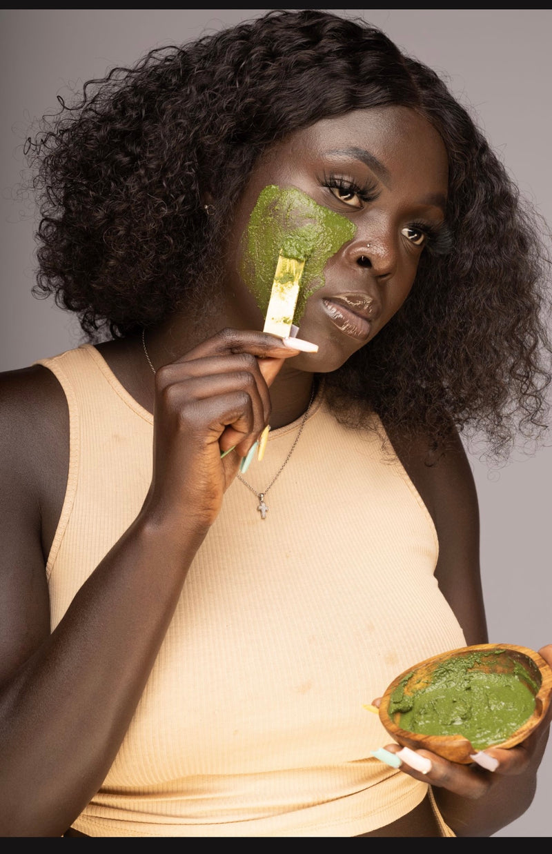 A person applying a green face scrub to their cheek with a brush, holding a bowl of the scrub in their other hand.